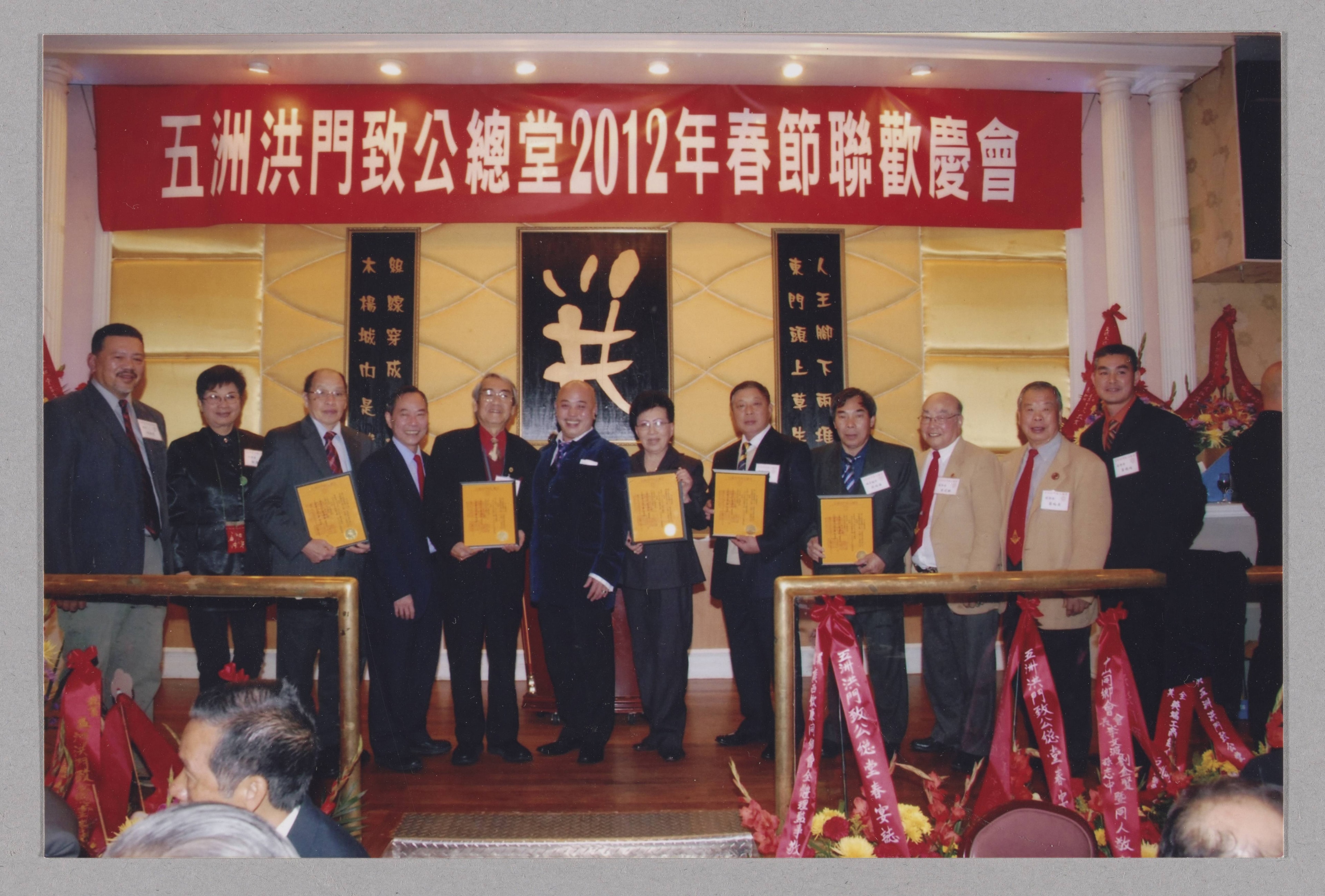 Supreme Lodge Lunar New Year celebration (五洲洪門致公總堂2012年春節聯歡慶會). Group holding commemorative plaques beneath celebration banner. This is an archival photograph of a past event; individuals pictured are not identified.