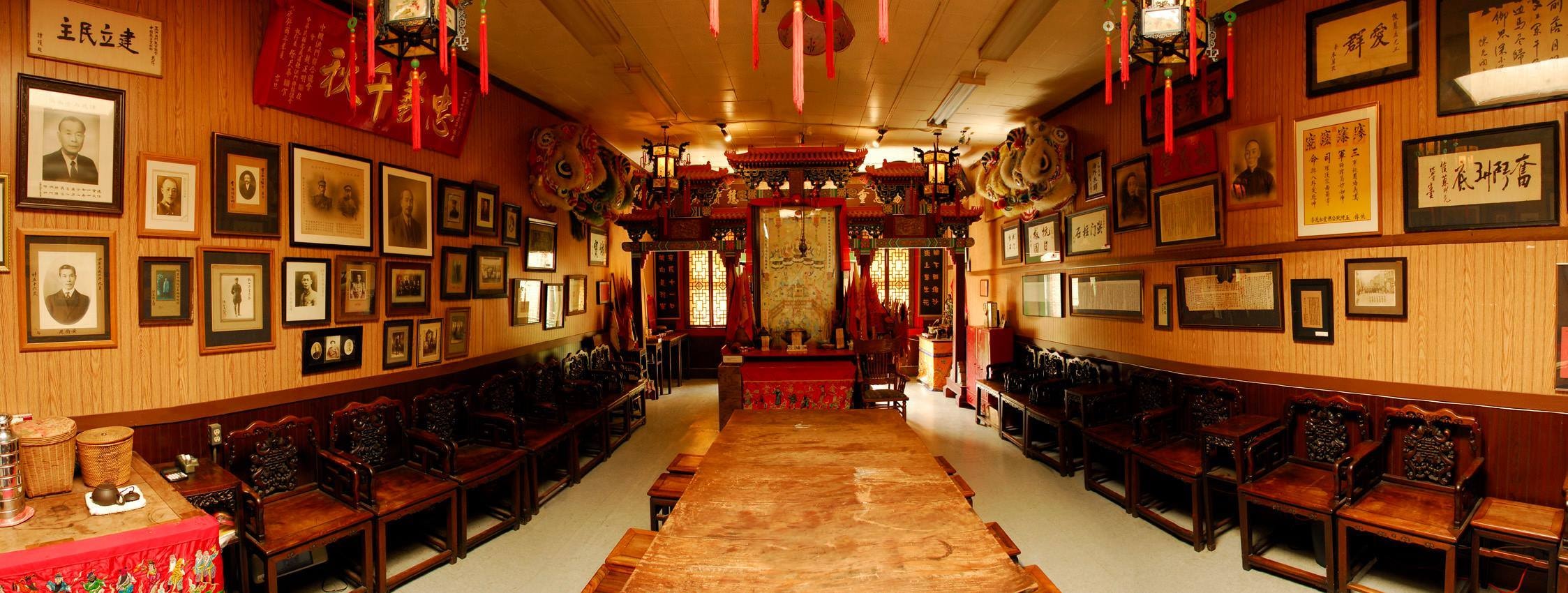 Interior of Chee Kung Tong's San Francisco headquarters, showing the meeting table, carved wooden seating, framed historical portraits and ceremonial altar.